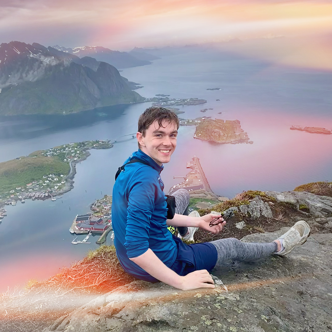 public affairs professional sitting on a rock at the edge of a cliff and smiling. View of water and mountains behind him.
