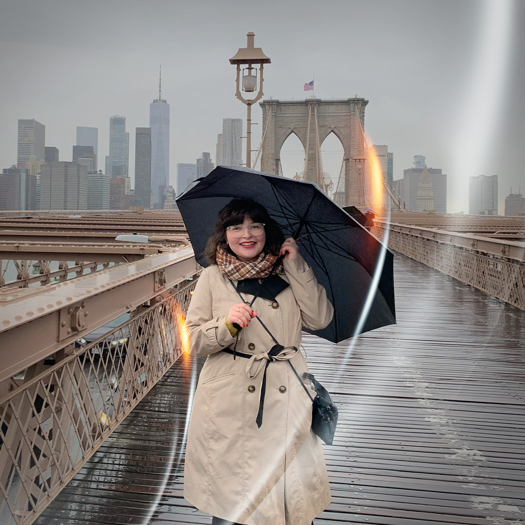 Digital marketing professional stood on Brooklyn Bridge wearing a trench coat and holding an umbrella with a smile.