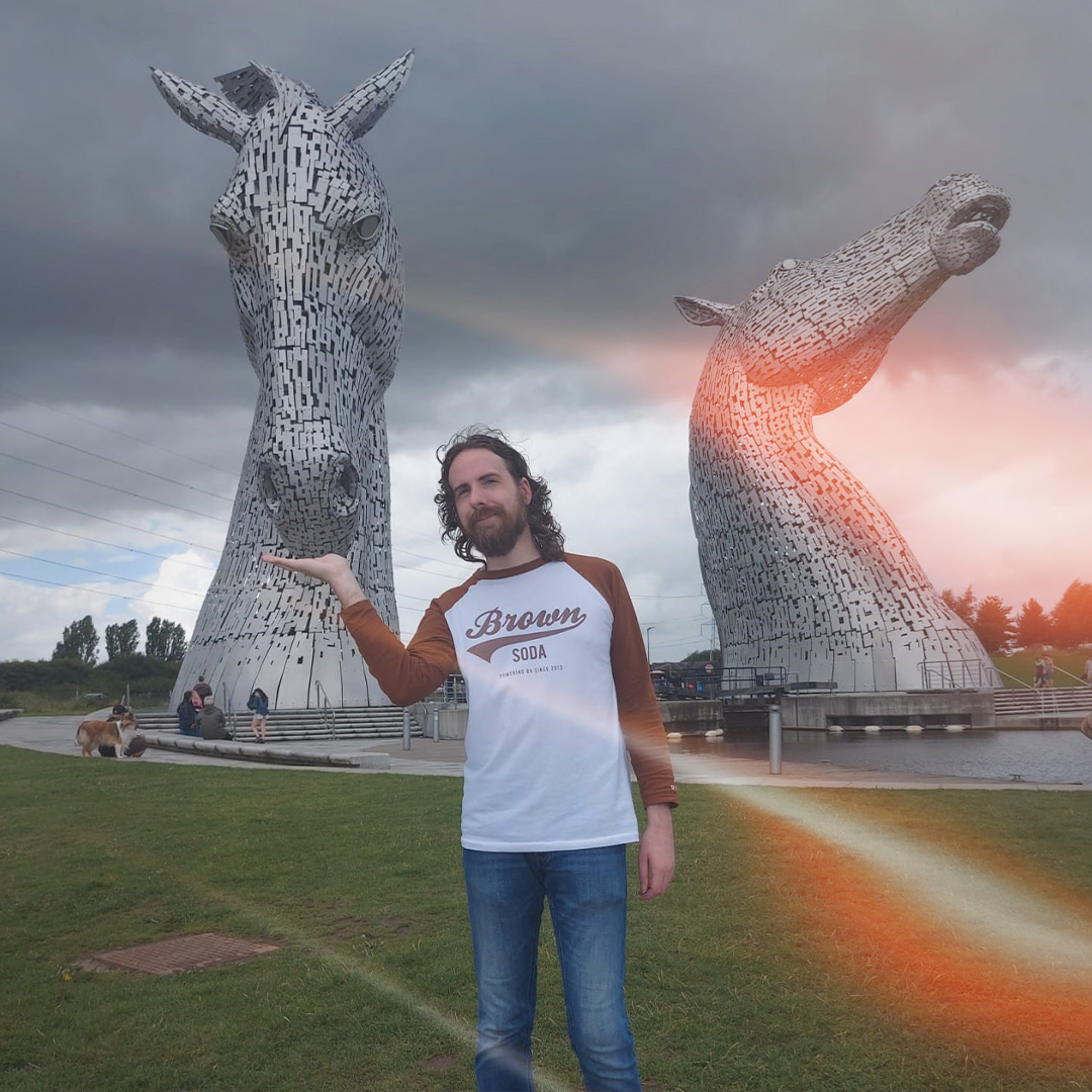 Man stood in front of The Kelpies holding his hand out as though one of them is eating out of it.